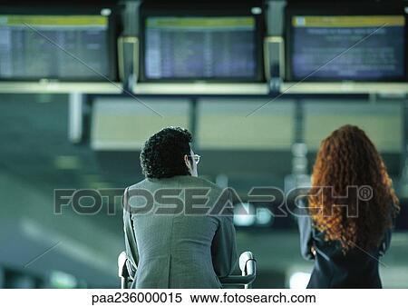Stock Photography - Businessman and businesswoman sitting under airport departure and arrival monitors, rear view. Fotosearch