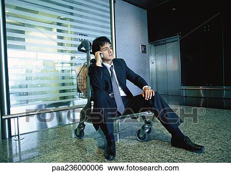 Businessman sitting on luggage cart talking on cell phone. View Large Photo Image Stock Photograph - Businessman sitting on luggage cart talking on cell phone. . Fotosearch