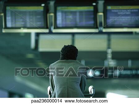 Picture - Businessman sitting under airport departure and arrival monitors, rear view. Fotosearch