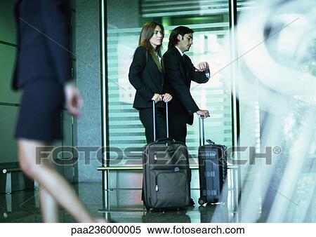 Stock Photography - Man and woman standing with suitcases, man looking at watch.. Fotosearch