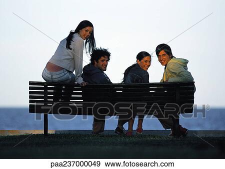 Stock Photo - Friends sitting on bench at sunset, smiling at camera.. Fotosearch