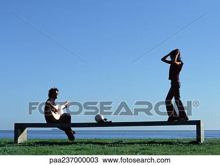 Stock Image - Man playing guitar, woman standing on bench near water. Fotosearch
