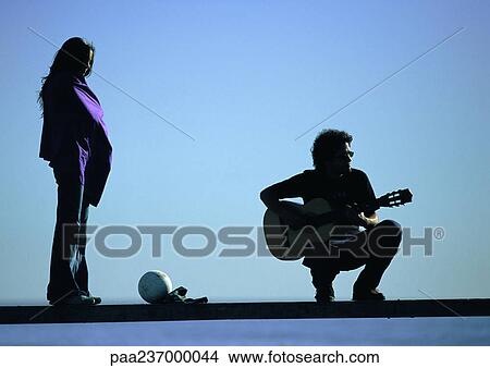 Woman standing, man squatting, playing guitar on bench. View Large Photo Image Picture - Woman standing, man squatting, playing guitar on bench.. Fotosearch