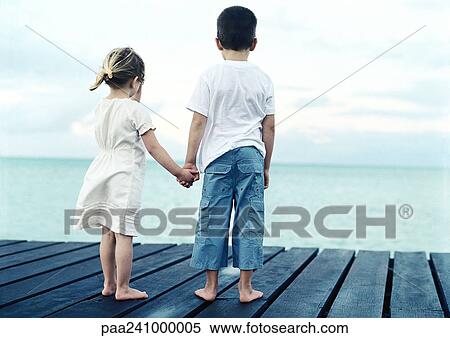 Stock Photography - Boy and girl standing on dock holding hands, rear view. Fotosearch