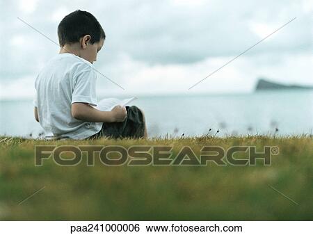 Stock Photograph - Boy reading on grass, sea in background, rear view. Fotosearch