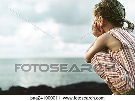 Stock Image - Girl crouching, sea in background, rear view. Fotosearch