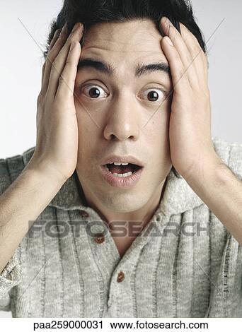 Stock Image - Young man looking at camera with eyes wide open and eyebrows raised, holding face with hands. Fotosearch