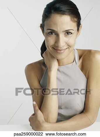 Stock Image - Young woman smiling at camera with one arm in front of chest and one hand holding neck. Fotosearch