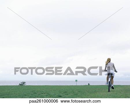 Woman riding bicycle on grass, sea and overcast sky in background View Large Photo Image Stock Photograph - Woman riding bicycle on grass, sea and overcast sky in background. Fotosearch