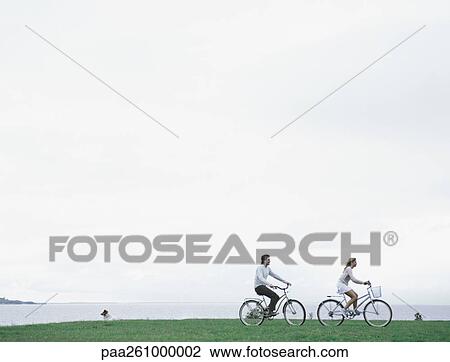 Young man and woman bicycling on grass with sea and overcast sky in background View Large Photo Image Stock Image - Young man and woman bicycling on grass with sea and overcast sky in background. Fotosearch