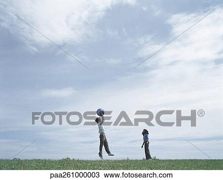 Stock Image - Young man and woman on grass in distance playing with ball, man jumping to catch ball. Fotosearch
