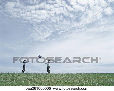 Stock Photography - Young man and woman standing on grass in distance, throwing ball. Fotosearch