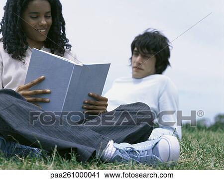 Young woman sitting on grass reading book, young man leaning on elbow behind her View Large Photo Image Stock Image - Young woman sitting on grass reading book, young man leaning on elbow behind her. Fotosearch