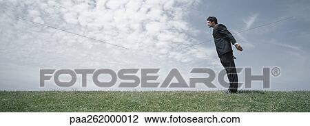 Stock Image - Man in suit standing on grass leaning forward with arms lifted backwards, side view. Fotosearch