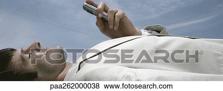 Stock Photo - Man lying on back holding cell phone with sky in background, close-up. Fotosearch
