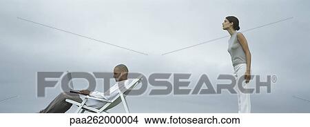 Picture - Man sitting in lounge chair with laptop on lap, woman standing peering over man's shoulder, side view. Fotosearch