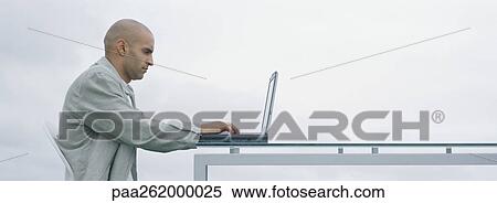 Stock Photography - Man sitting outdoors at table using laptop, side view. Fotosearch