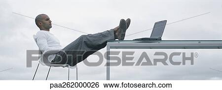 Stock Photograph - Man sitting outdoors with feet up on table and laptop on table, sideview. Fotosearch