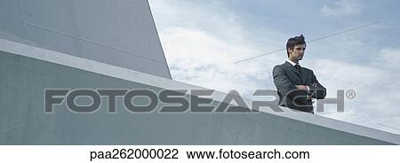 Stock Image - Man standing behind concrete wall with arms folded, low angle view. Fotosearch
