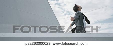 Stock Image - Man standing behind concrete wall with arms outstretched, side view. Fotosearch