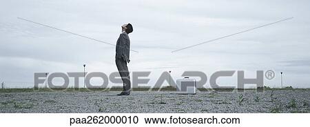 Stock Image - Man standing in abandoned lot looking up at sky, metallic briefcase on ground behind him. Fotosearch