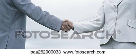 Stock Image - Woman and man in suits shaking hands, mid-section with sky in background. Fotosearch