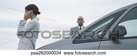 Picture - Woman and man in suits standing near car, man looking at camera, woman talking on cell phone. Fotosearch