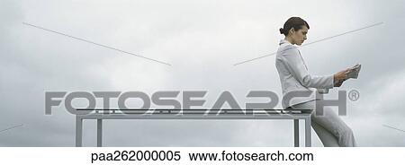 Stock Photography - Woman leaning against edge of table, holding newspaper, side view, in front of overcast sky. Fotosearch