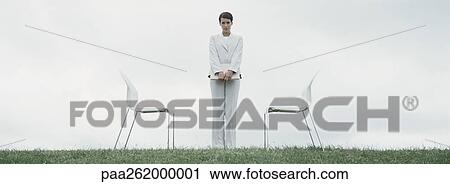 Stock Image - Woman standing outdoors on grass between two chairs in front of overcast sky. Fotosearch