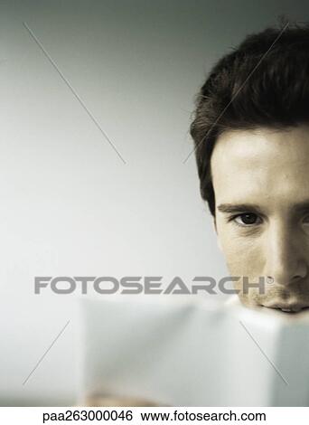 Stock Photograph - Man looking at camera from behind open book, partial view. Fotosearch