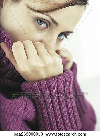 Stock Image - Woman covering part of face with neck of sweater, looking at camera, close-up. Fotosearch