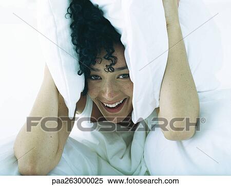 Stock Photography - Woman in bed holding pillows to both sides of head, looking at camera. Fotosearch