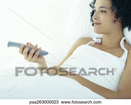 Stock Image - Woman in bed under covers pointing remote control. Fotosearch