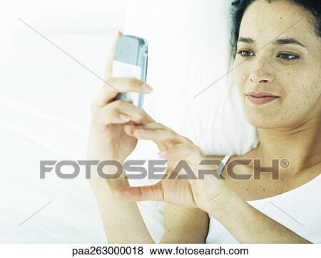 Stock Photo - Woman lying on back on bed, looking at cell phone. Fotosearch
