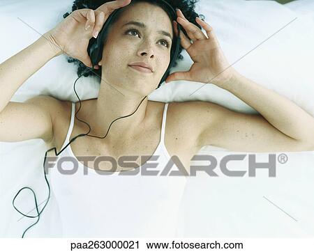 Stock Image - Woman lying on back on bed with earphones on and hands at side of head. Fotosearch