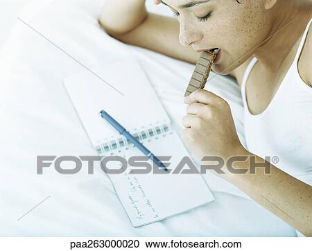 Woman lying on side on bed eating chocolate and looking down at notebook with pen lying across it View Large Photo Image Stock Image - Woman lying on side on bed eating chocolate and looking down at notebook with pen lying across it. Fotosearch