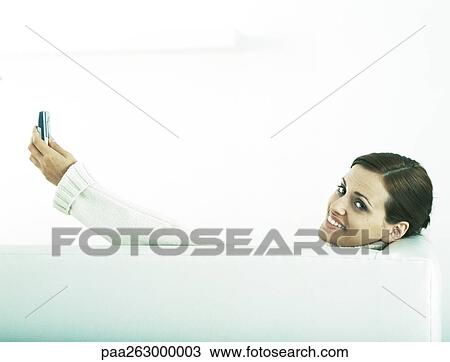 Stock Image - Woman sitting on couch, rear view, looking back at camera and holding up cell phone. Fotosearch