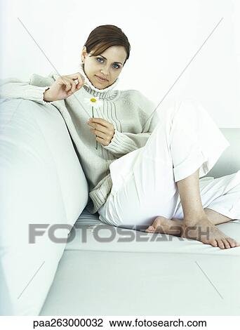 Stock Image - Woman sitting with knee up on sofa, plucking petals from daisy. Fotosearch