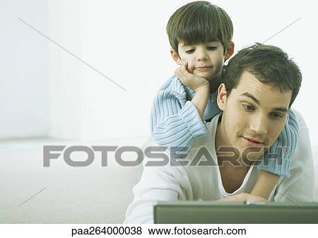 Stock Photo - Little boy on man's back, both looking down at laptop. Fotosearch