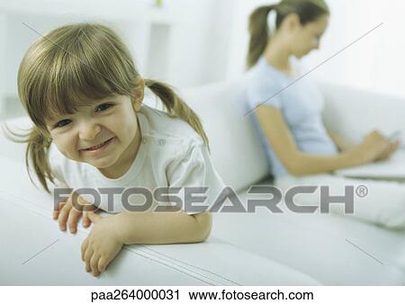 Stock Image - Little girl leaning on sofa arm making faces at camera, young woman looking down in background. Fotosearch