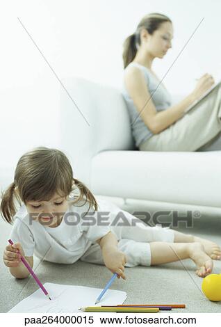 Stock Photography - Little girl lying on floor coloring, young woman sitting behind her on couch writing with knees up. Fotosearch