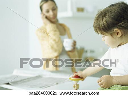 Stock Photography - Little girl playing with toy, woman holding mug and talking on phone in background. Fotosearch