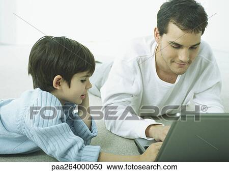 Stock Image - Man and little boy lying on floor on stomachs, working on laptop. Fotosearch