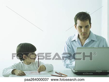 Stock Photo - Man sitting at table working on laptop, boy sitting next to him coloring with pencils. Fotosearch