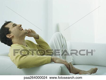 Picture - Man sitting barefoot on couch talking on phone, laughing with head back. Fotosearch