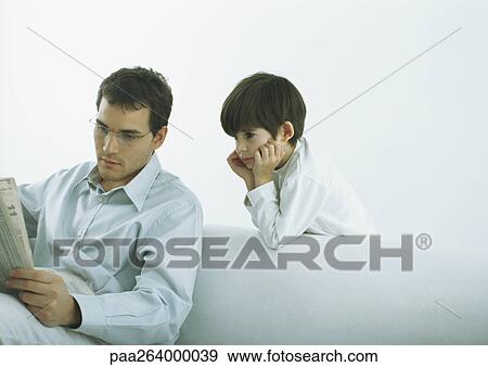 Stock Photo - Man sitting on sofa reading newspaper, boy standing behind sofa looking over man's shoulder. Fotosearch