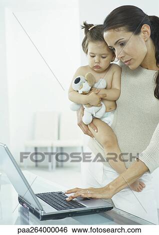 Stock Photograph - Woman holding little girl in one arm, typing on laptop with other hand. Fotosearch