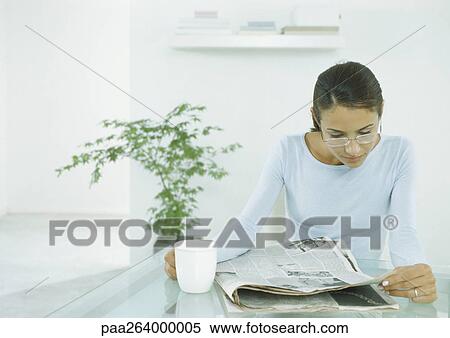 Stock Photography - Woman sitting at table holding mug and looking at newspaper. Fotosearch