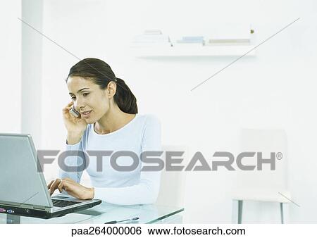 Woman sitting at table typing on laptop, holding cell phone to ear View Large Photo Image Stock Photograph - Woman sitting at table typing on laptop, holding cell phone to ear. Fotosearch