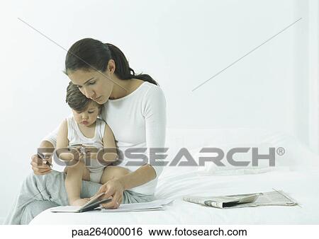 Woman sitting on edge of bed with little girl on lap, looking down at agenda View Large Photo Image Stock Photograph - Woman sitting on edge of bed with little girl on lap, looking down at agenda. Fotosearch